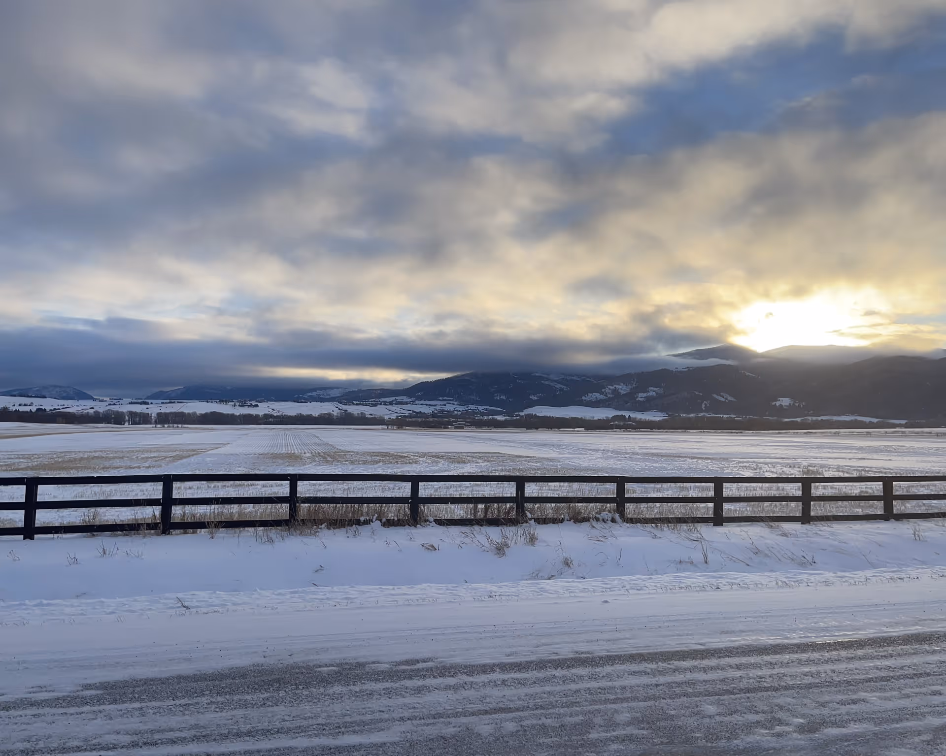 Snow-covered field with a wooden fence in the foreground and mountains in the background under a cloudy sky with the sun partially visible.