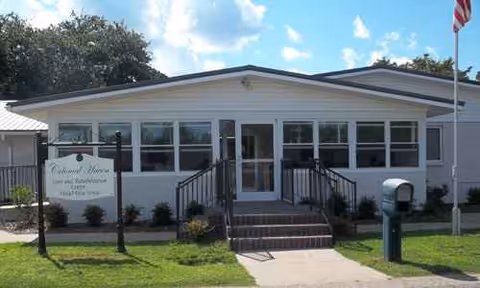 Single-story care facility with front entrance, steps, sign, flag, and manicured lawn.