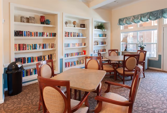 A cozy common area with several wooden tables and cushioned chairs arranged on a patterned carpet. The room features built-in white bookshelves filled with books and decorative items, a large window with a green valance, and a lamp on a side table near the window. A black trash can is visible near the bookshelves.