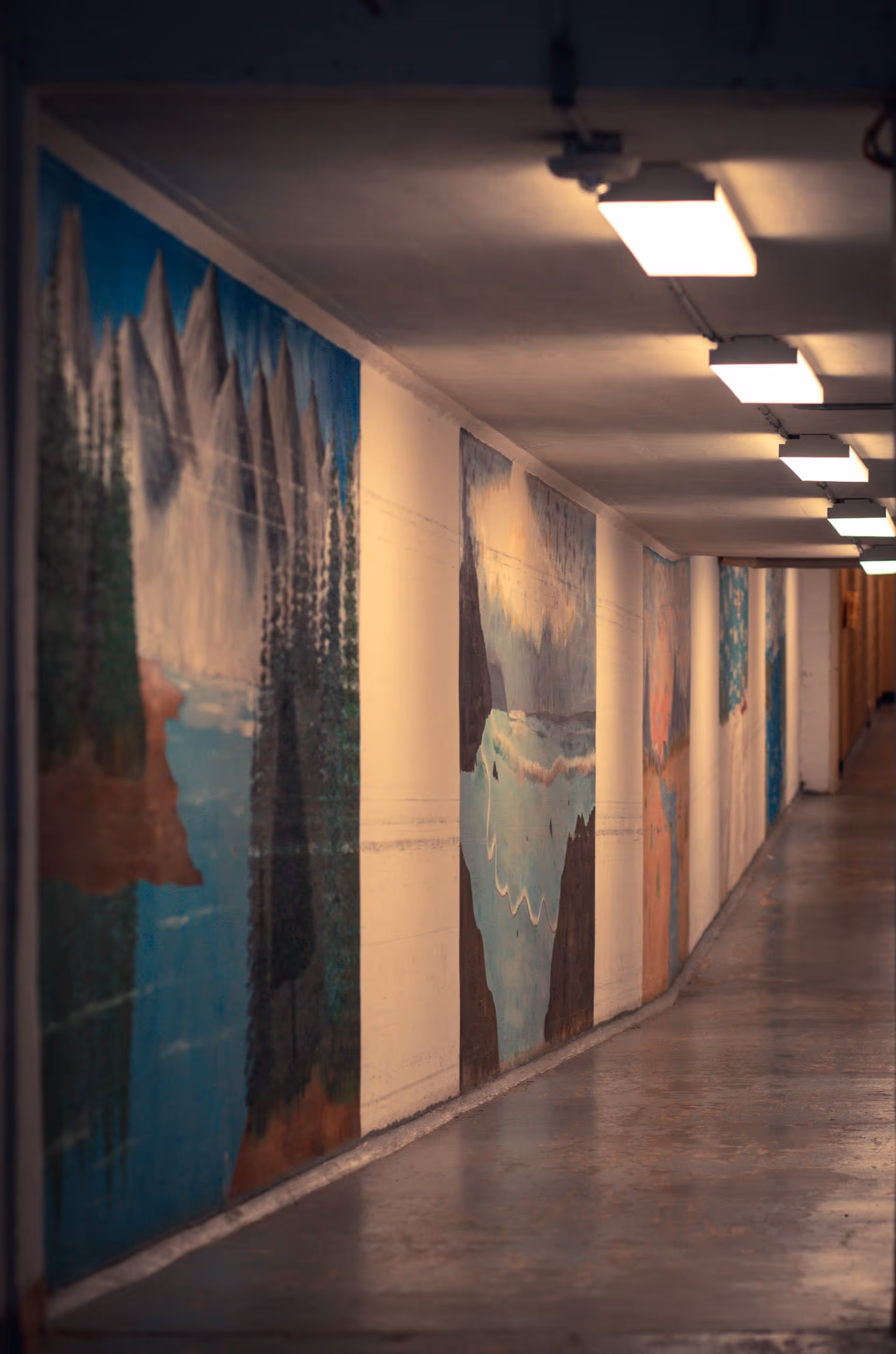 A long indoor hallway with a series of colorful murals depicting nature scenes such as mountains, trees, and water on the left wall. The hallway is lit by rectangular ceiling lights and has a polished concrete floor.