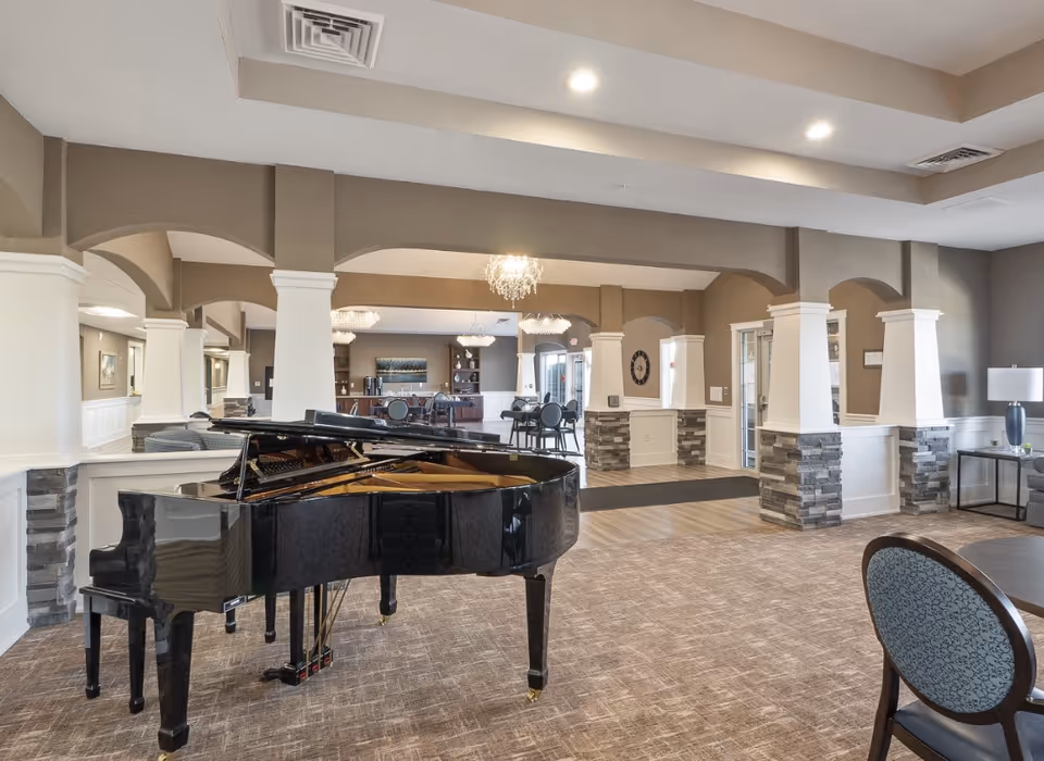 Spacious and elegant common area in a senior living facility featuring a black grand piano in the foreground, multiple seating areas with chairs and tables, decorative columns with stone accents, and modern chandeliers hanging from the ceiling.