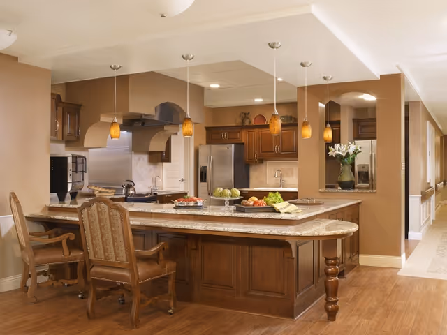 A spacious kitchen area with a large granite countertop island featuring wooden cabinetry. The island has two upholstered wooden chairs and is decorated with plates of food and a bowl of fruit. Above the island, there are five pendant lights with amber-colored shades. The kitchen includes stainless steel appliances, including a refrigerator and a range hood. The walls are painted in warm beige tones, and there is a hallway visible to the right.