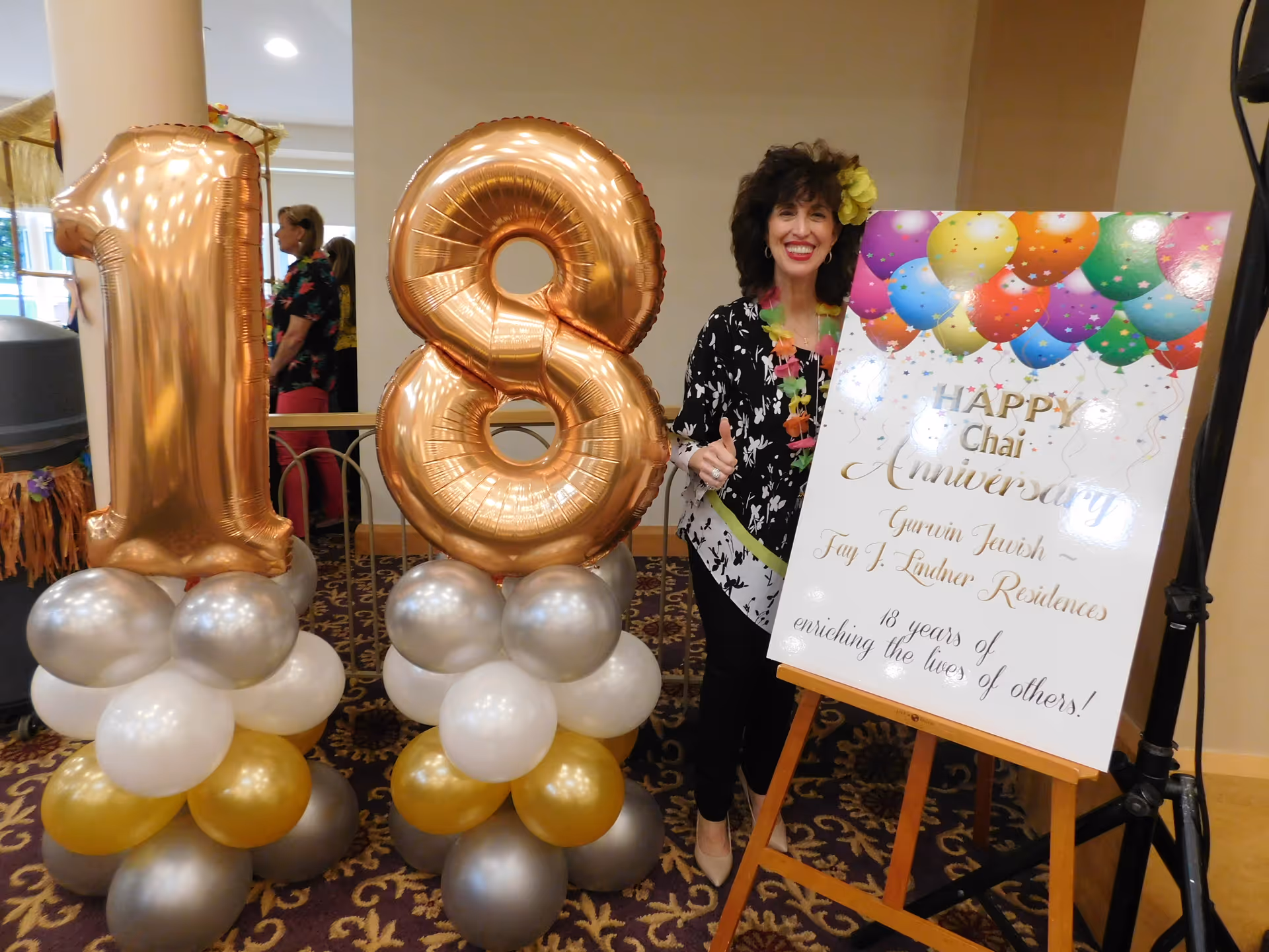 A woman standing next to large gold balloons shaped as the number 18 and a colorful sign celebrating the 18th anniversary of Gurwin Jewish - Fay J. Lindner Residences. The sign has balloons printed on it and reads 'HAPPY Chai Anniversary Gurwin Jewish - Fay J. Lindner Residences 18 years of enriching the lives of others!'. The woman is smiling and giving a thumbs up.