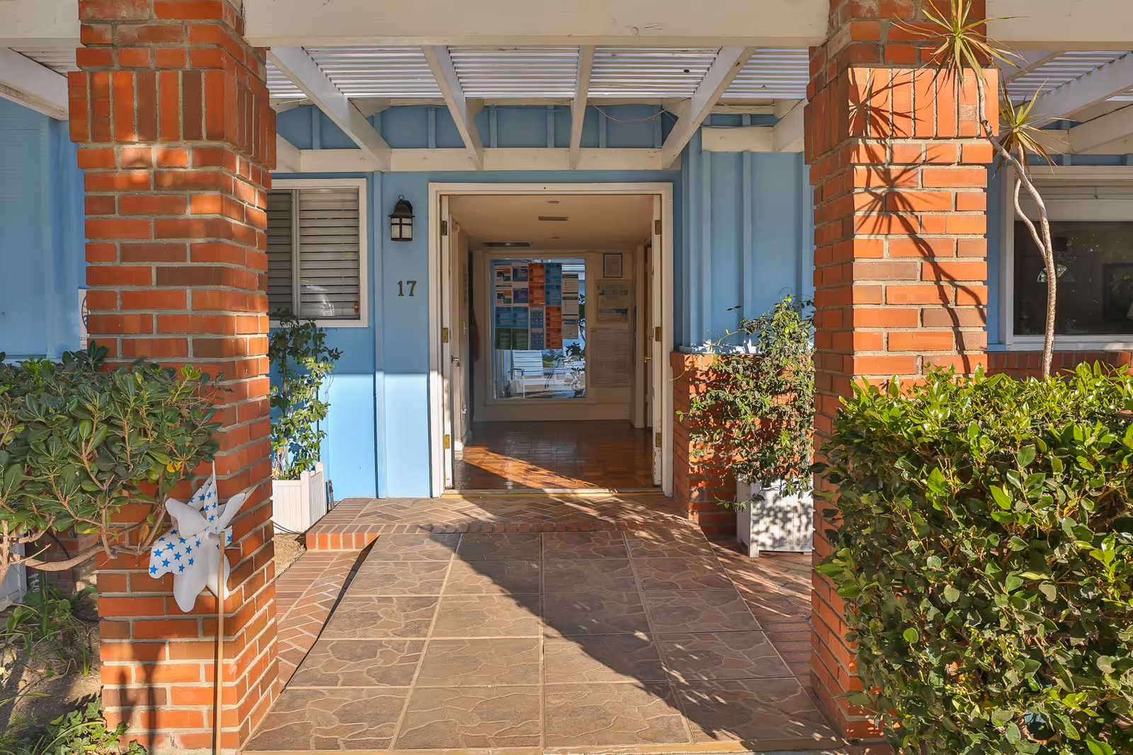 Entrance to a light-blue senior living building framed by brick columns, potted plants, and a tiled walkway leading to an open door marked 17.