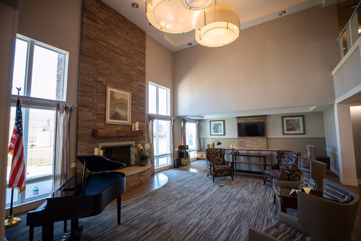 A spacious living room with high ceilings featuring a stone fireplace, a grand piano, several patterned armchairs, a TV mounted on a stone accent wall, large windows with curtains, and modern hanging light fixtures.