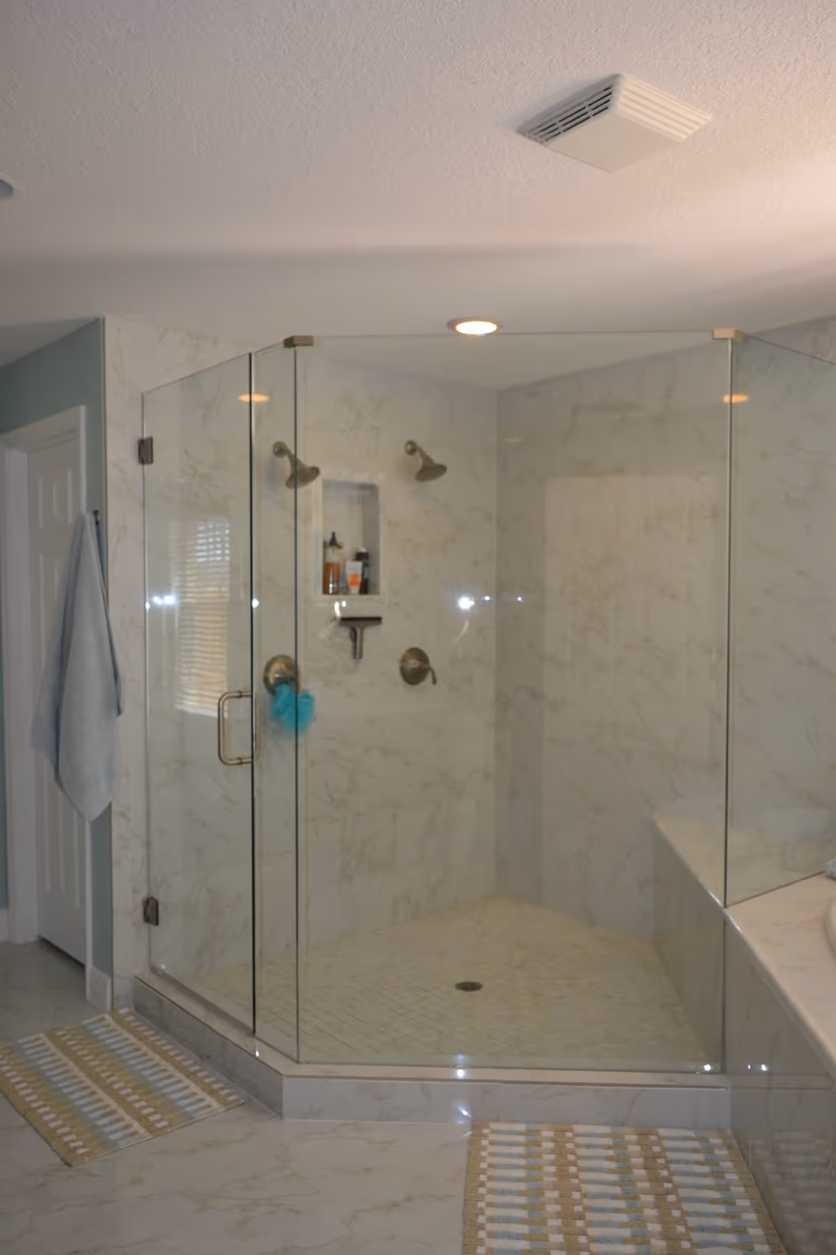 A modern bathroom featuring a large glass-enclosed corner shower with two shower heads and marble-patterned walls and floor. There are two striped bath mats on the floor and a towel hanging on a hook next to a white door.