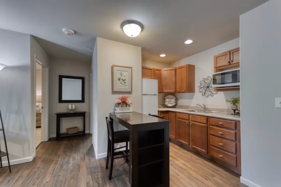 Interior view of a kitchen area in a senior living facility with wooden cabinets, a white refrigerator, a microwave, a sink, and a small dining table with two chairs. The floor is wood, and there is a small table with candles and a mirror in the hallway leading to another room.