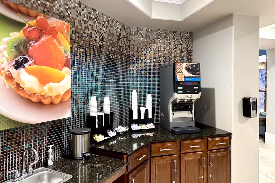 A coffee and beverage station with a drink dispenser, stacked disposable cups, sink and countertop against a mosaic tile backsplash.