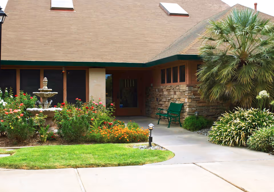 Outdoor view of a senior living facility entrance with a stone and wood exterior, a green bench, a small garden with colorful flowers, a multi-tiered stone fountain, and various shrubs and palm plants along a concrete pathway.
