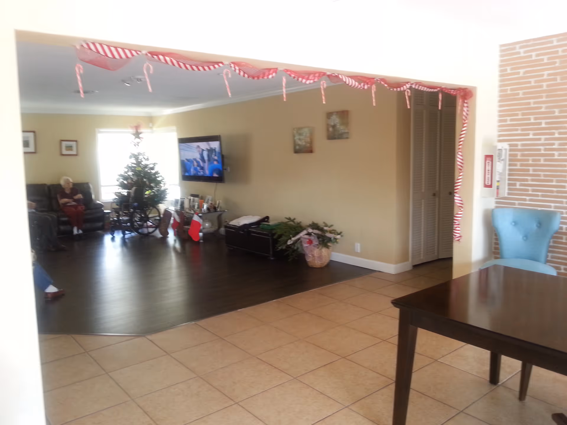 Interior view of a senior living facility common area decorated for Christmas with candy cane ribbons hanging from the ceiling. There is a Christmas tree with stockings and a wheelchair nearby. An elderly person is sitting on a couch, and a TV is mounted on the wall. The floor transitions from tile to dark wood, and there is a blue chair and a dark wooden table in the foreground.