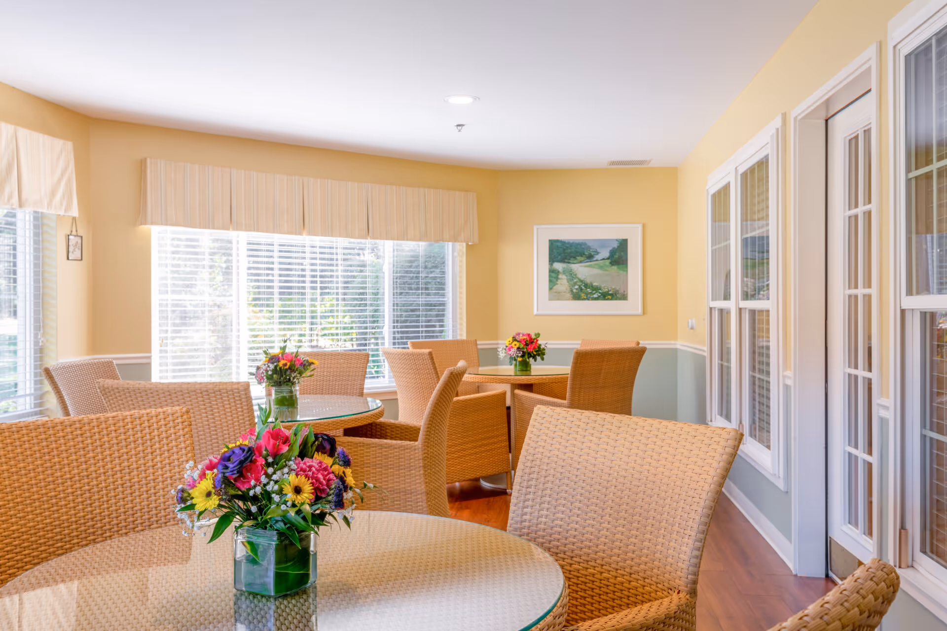 Bright dining area with wicker chairs and round glass-topped tables arranged near large windows and floral centerpieces.