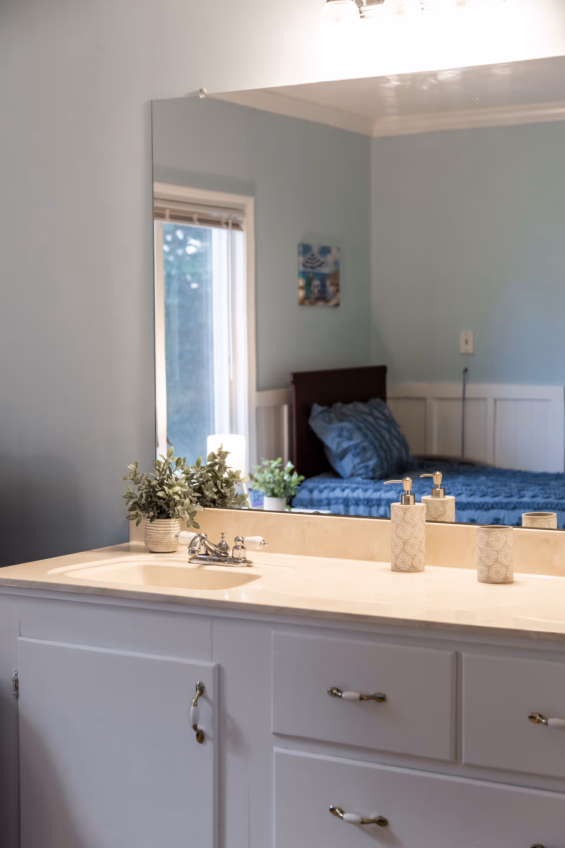 A bathroom sink countertop with a faucet, soap dispenser, and a small potted plant. A large mirror above the sink reflects a bedroom with a bed covered in blue bedding and a window with blinds.
