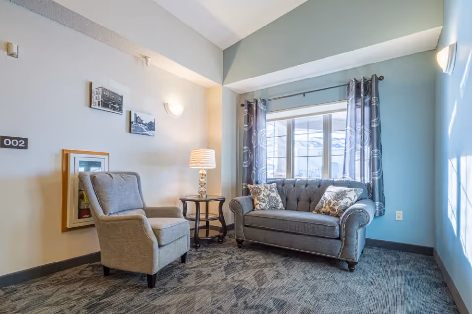 A cozy sitting area in a senior living facility with a gray upholstered armchair and a matching gray loveseat adorned with patterned throw pillows. A round side table with a decorative lamp is placed between the chairs. The room has light blue walls, a large window with patterned curtains, and carpeted flooring. Two black and white framed photos hang on the wall above the armchair.