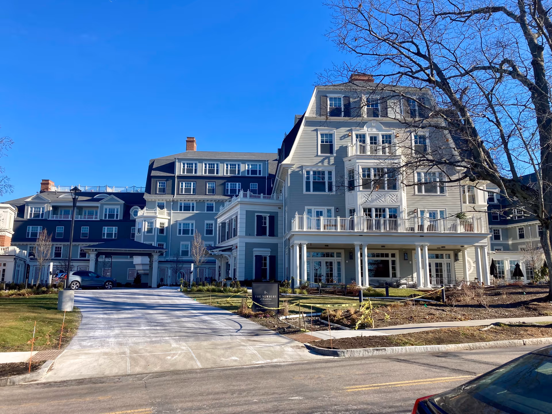 Exterior view of The Newbury Of Brookline, a large multi-story senior living facility with light gray siding, white trim, and multiple windows. The building has a driveway leading to a covered entrance, landscaped grounds, and a clear blue sky overhead.