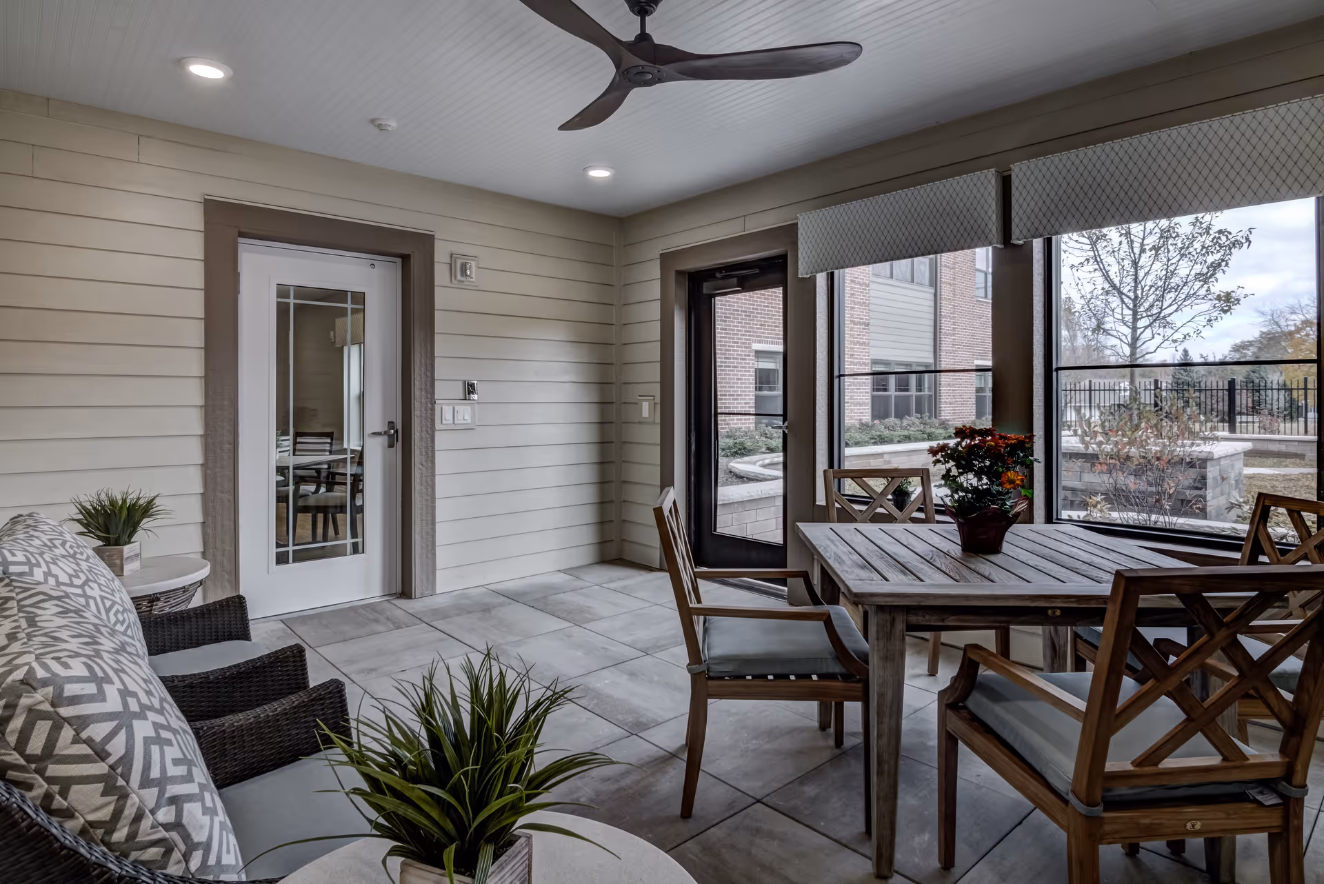 A cozy indoor seating area with a wooden table and four cushioned chairs, a wicker sofa with patterned cushions, potted plants, and large windows showing an outdoor garden area with a fire pit. The ceiling has recessed lighting and a modern ceiling fan.