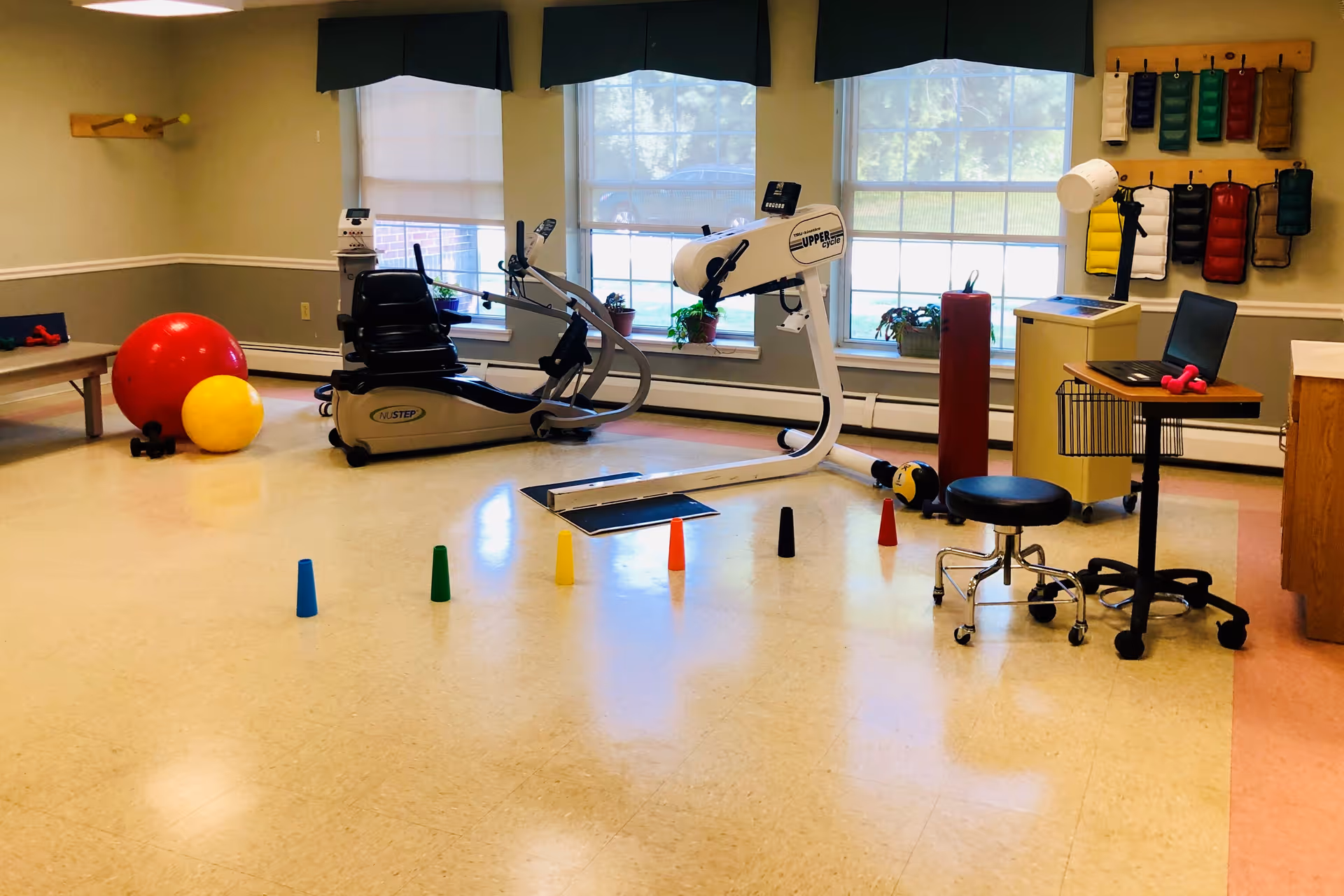 A physical therapy or exercise room with large windows, exercise equipment including a recumbent bike and an upper body ergometer, colorful cones on the floor, large red and yellow exercise balls, small dumbbells, and a desk with a laptop and lamp.