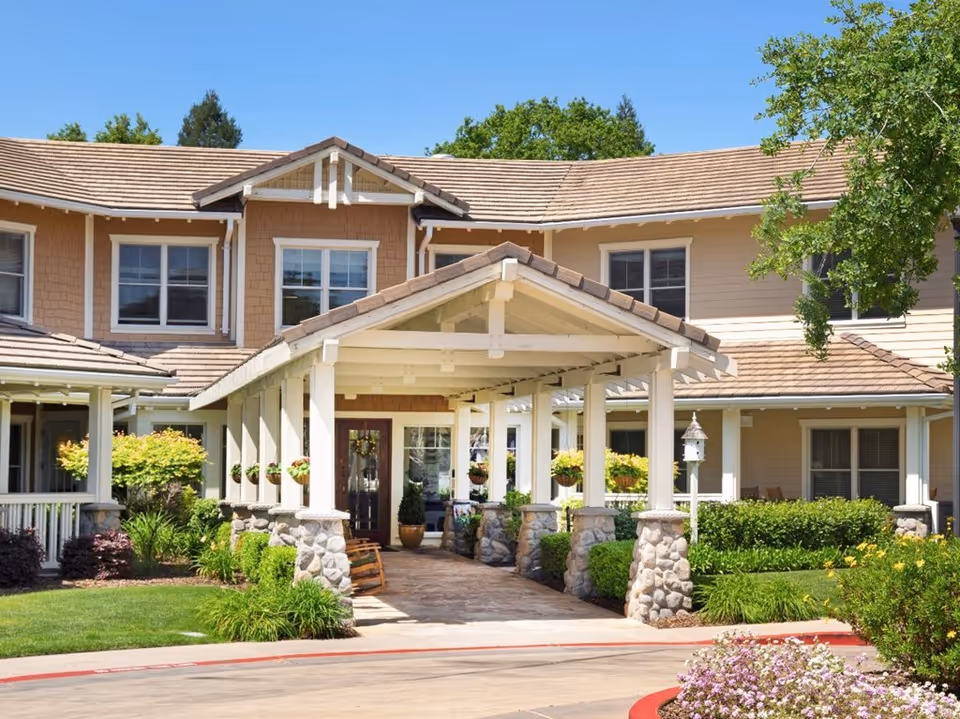 Front entrance of a two-story senior living building with a covered porte-cochère supported by stone pillars, hanging plants, and landscaped grounds.