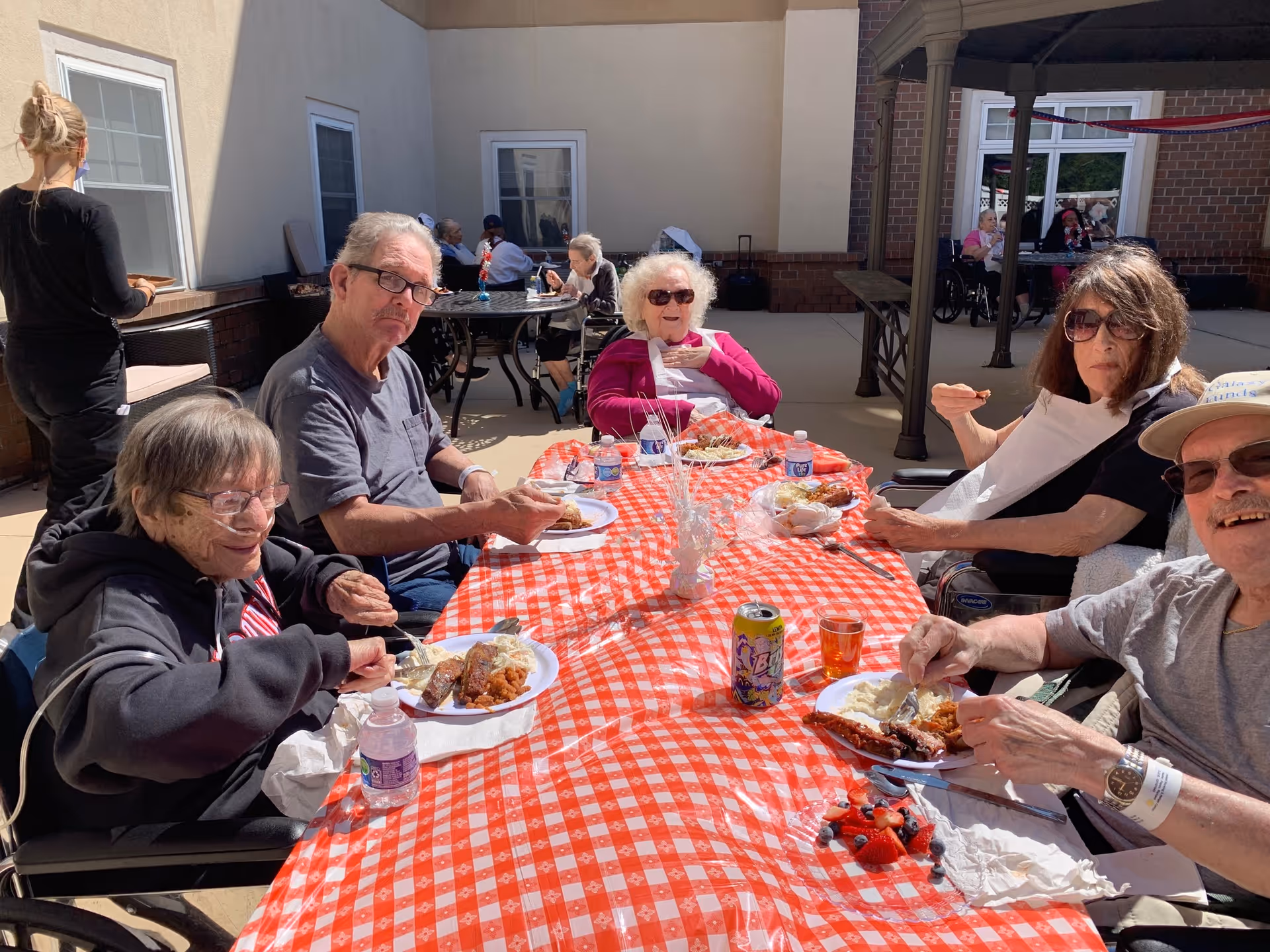 Several older adults sit around a red-and-white checkered outdoor tablecloth eating meals on paper plates in a courtyard.