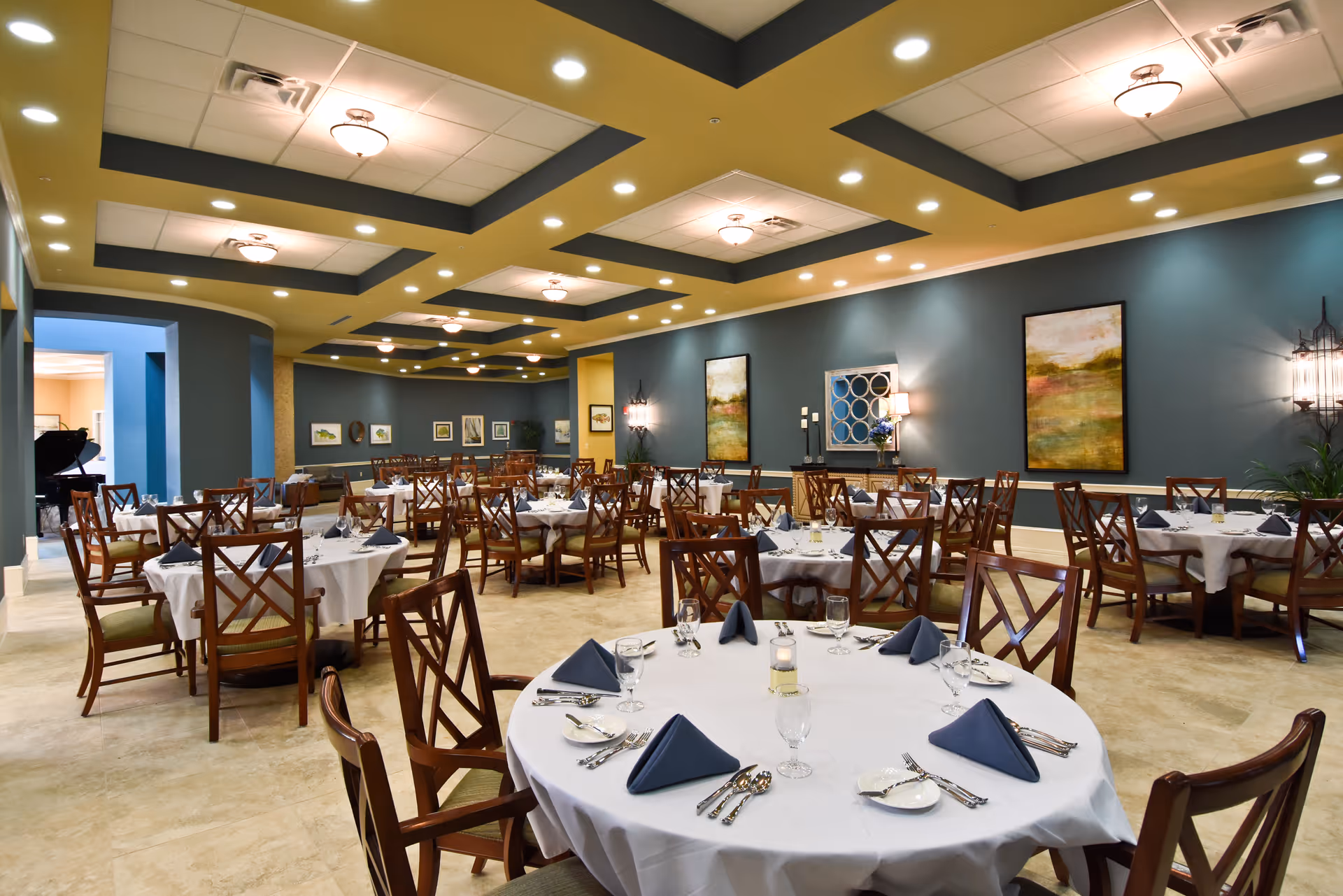 A spacious dining room with multiple round tables covered with white tablecloths, each set with blue folded napkins, glassware, plates, and silverware. The room has blue walls adorned with framed artwork and decorative lighting fixtures. The ceiling features recessed lighting and decorative panels.