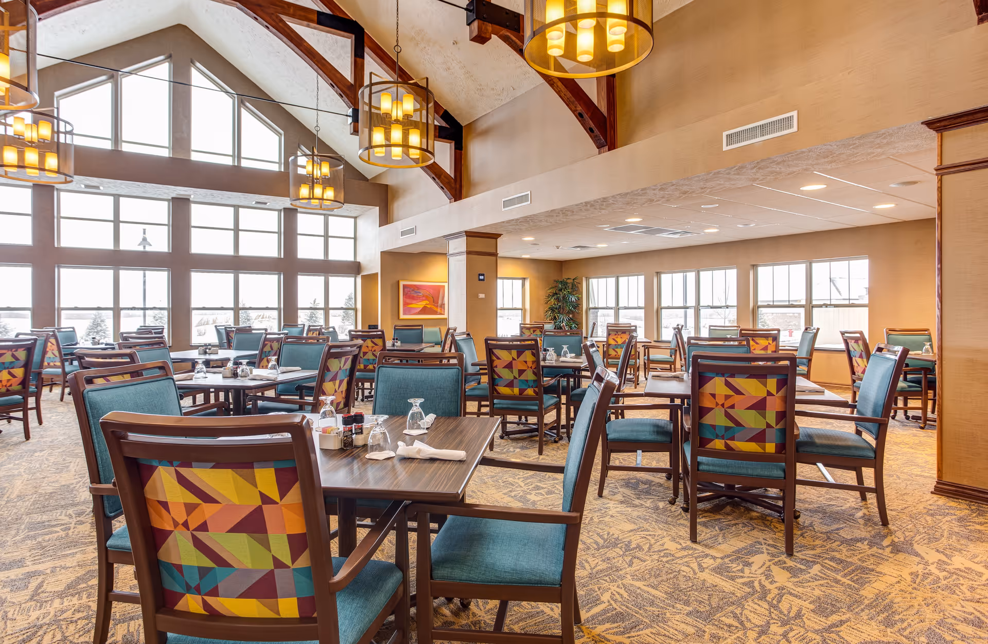 Bright dining room with multiple tables and colorful upholstered chairs beneath vaulted wooden beams and large windows.