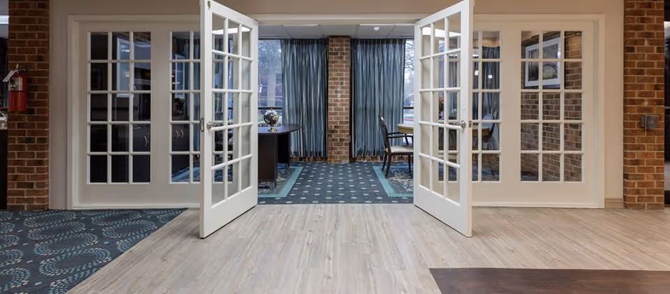 Interior view of a room with double glass-paneled doors open, revealing a carpeted area with a table and chairs near large windows with blue curtains. The room has brick walls and a mix of wood and carpet flooring.