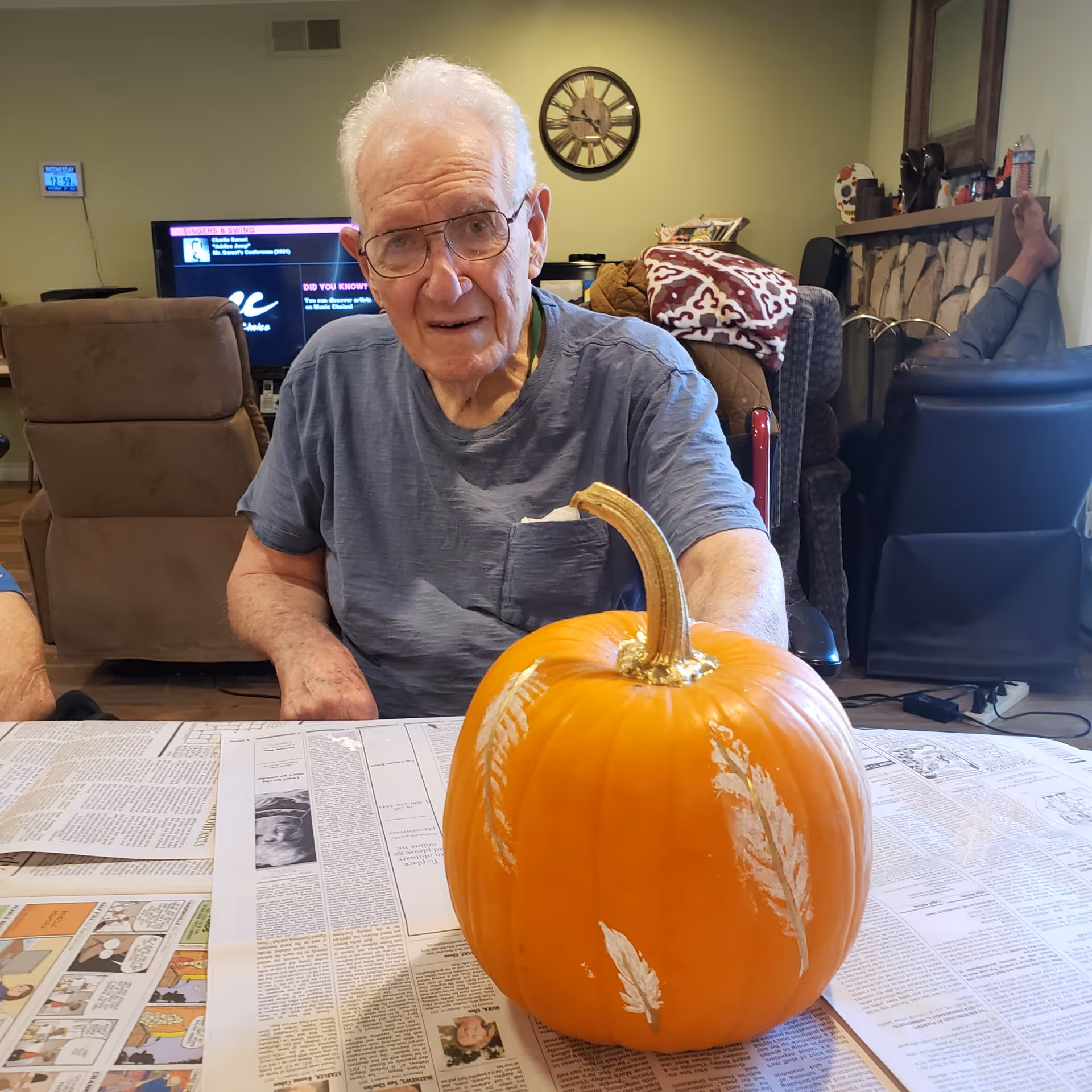 An elderly man wearing glasses and a blue shirt sits at a table covered with newspapers. In front of him is a medium-sized orange pumpkin decorated with white feather designs. Behind him is a living room with a brown recliner, a TV, a clock on the wall, and another person sitting with their feet up on a chair near a fireplace.