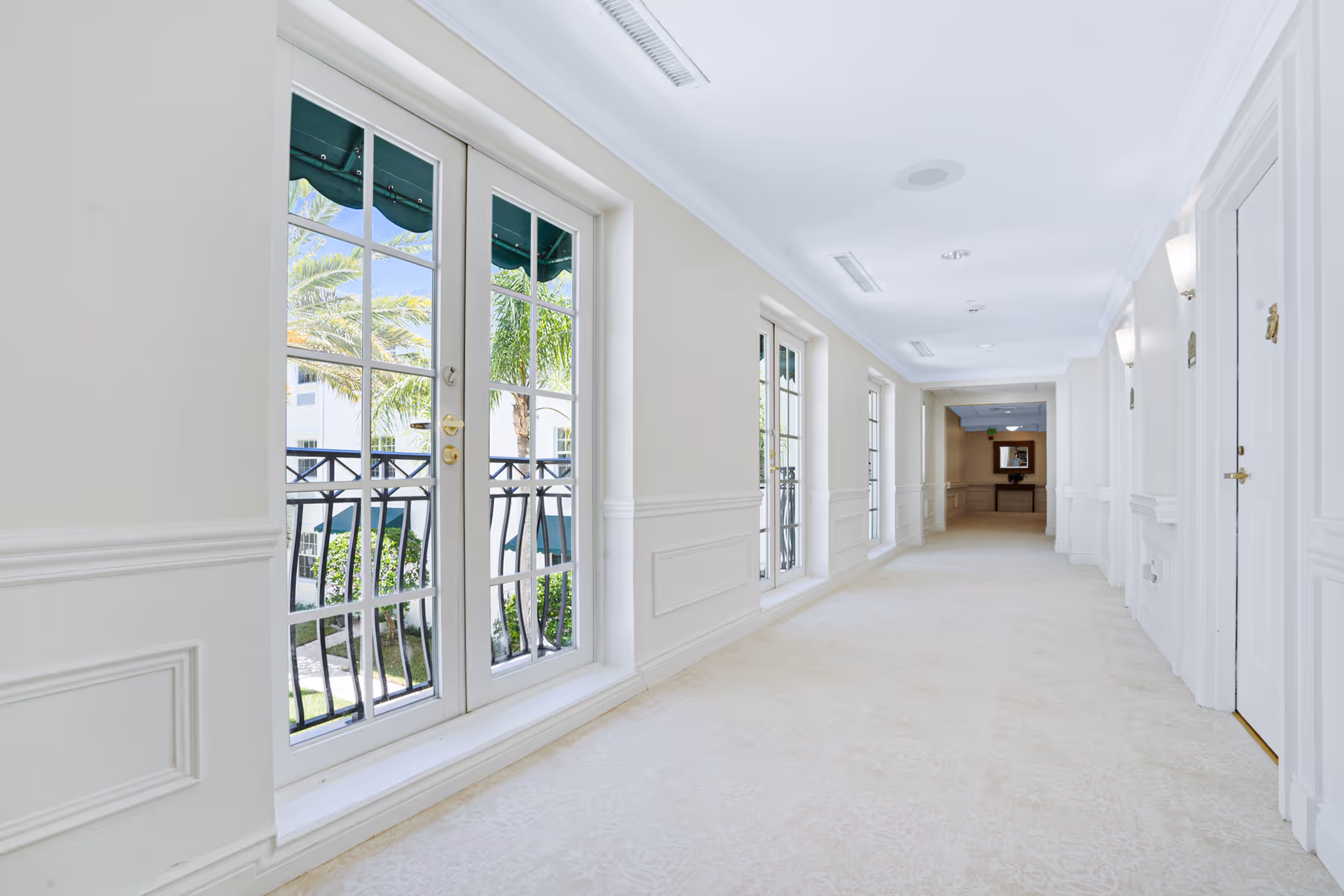 Bright and clean hallway in a senior living facility with large windows and glass doors on the left side showing palm trees outside, white walls with decorative molding, beige carpeted floor, and white doors on the right side with wall-mounted lights.