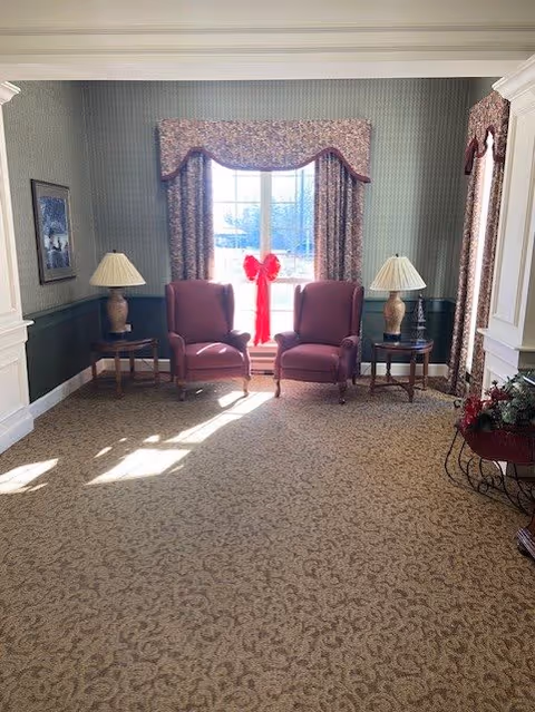 A cozy sitting area with two purple armchairs placed side by side in front of a window adorned with floral curtains and a red bow decoration. Each chair is flanked by a wooden side table with a lamp. The room has patterned carpet flooring, green and white walls, and a floral arrangement on the right side.