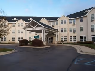 Exterior view of a multi-story senior living facility building with beige siding and numerous windows. The entrance features a covered drop-off area with the number 1140 displayed on the structure. There are bushes and a paved driveway in front of the building.