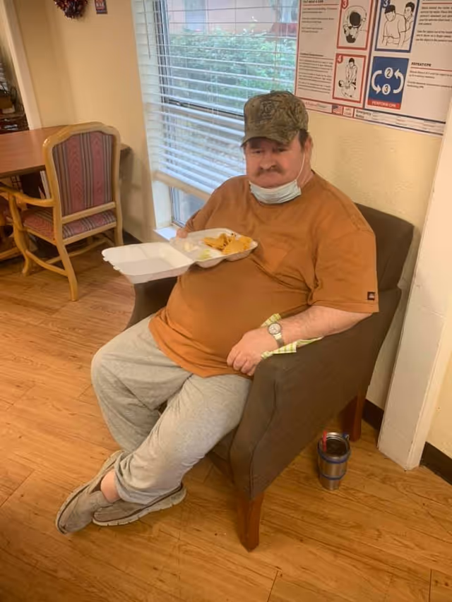A man wearing a brown shirt, gray sweatpants, and a camouflage cap is sitting in a brown armchair indoors. He is holding a takeout food container with some food in it. There is a window with blinds behind him and a poster with CPR instructions on the wall. A wooden floor and a table with chairs are visible in the background.
