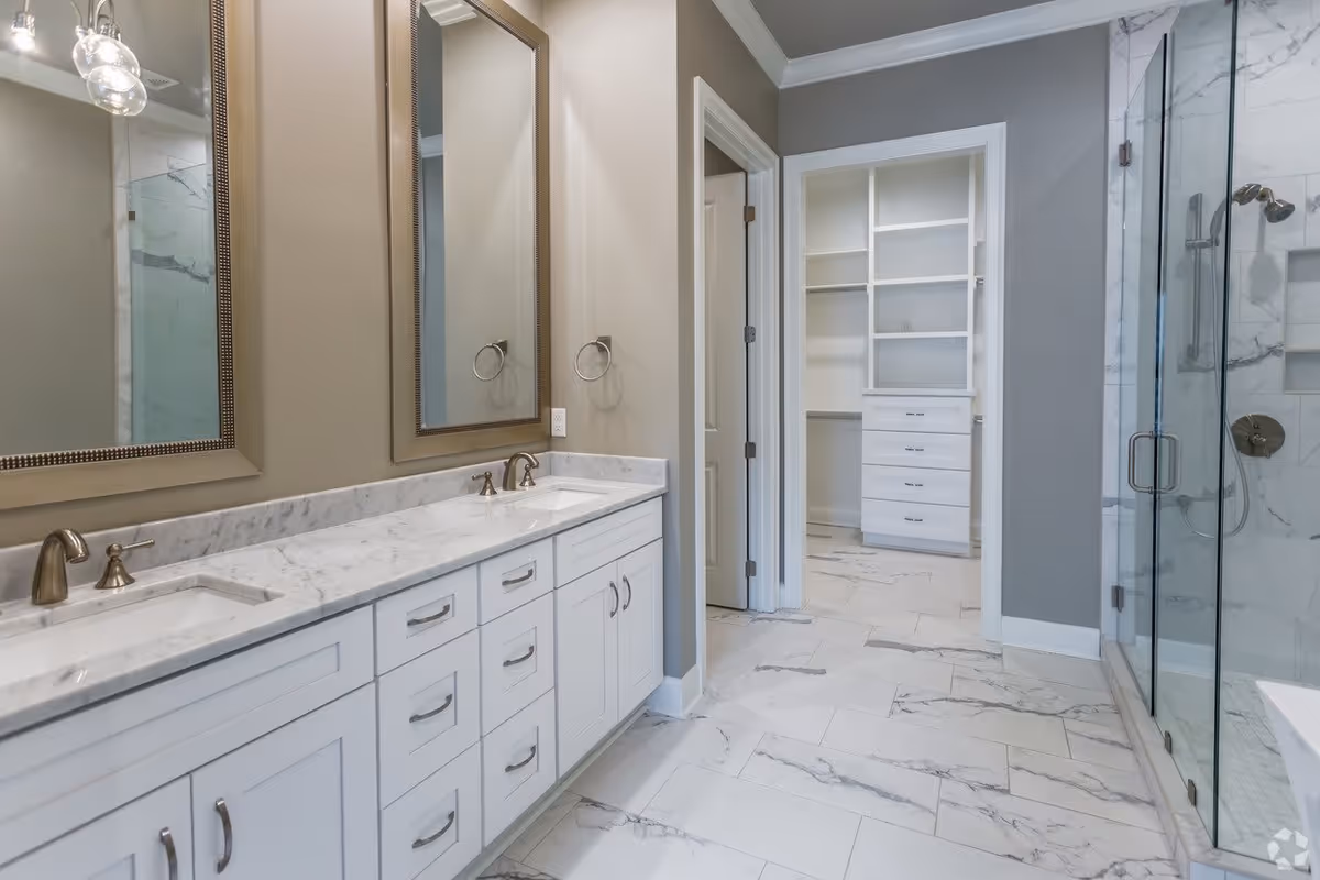 Modern bathroom with a double sink vanity featuring white cabinets and marble countertops, two large framed mirrors above the sinks, a glass-enclosed shower with marble tile walls, and an open doorway leading to a walk-in closet with built-in white shelving and drawers. The floor is covered with white marble tiles with gray veining.