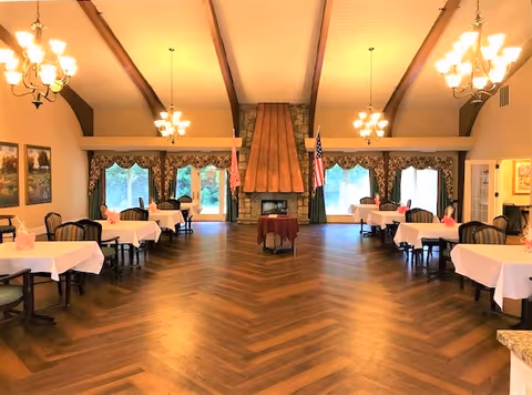 A spacious dining room with wooden herringbone flooring, several tables covered with white tablecloths and pink napkins, upholstered chairs, large windows with decorative curtains, a stone fireplace with a copper hood, and two chandeliers hanging from a vaulted ceiling with exposed wooden beams. Two flags are positioned on either side of the fireplace.