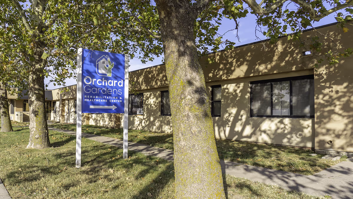 Exterior view of a single-story beige building with several windows, partially shaded by large trees. In front of the building is a blue sign that reads 'Orchard Gardens Rehabilitation & Healthcare Center' with an arrow pointing to the right.