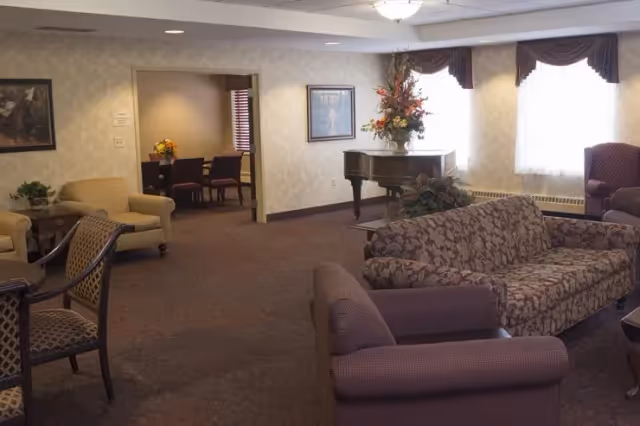 A cozy living room area in an assisted living facility featuring patterned sofas and armchairs arranged around a coffee table. There is a grand piano with a floral arrangement on top near two large windows with curtains. In the background, there is an adjoining dining area with a table and chairs, and framed artwork on the walls.
