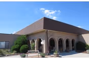 Front entrance of a brick building with arched columns, shrubs, planters, and a clear blue sky.