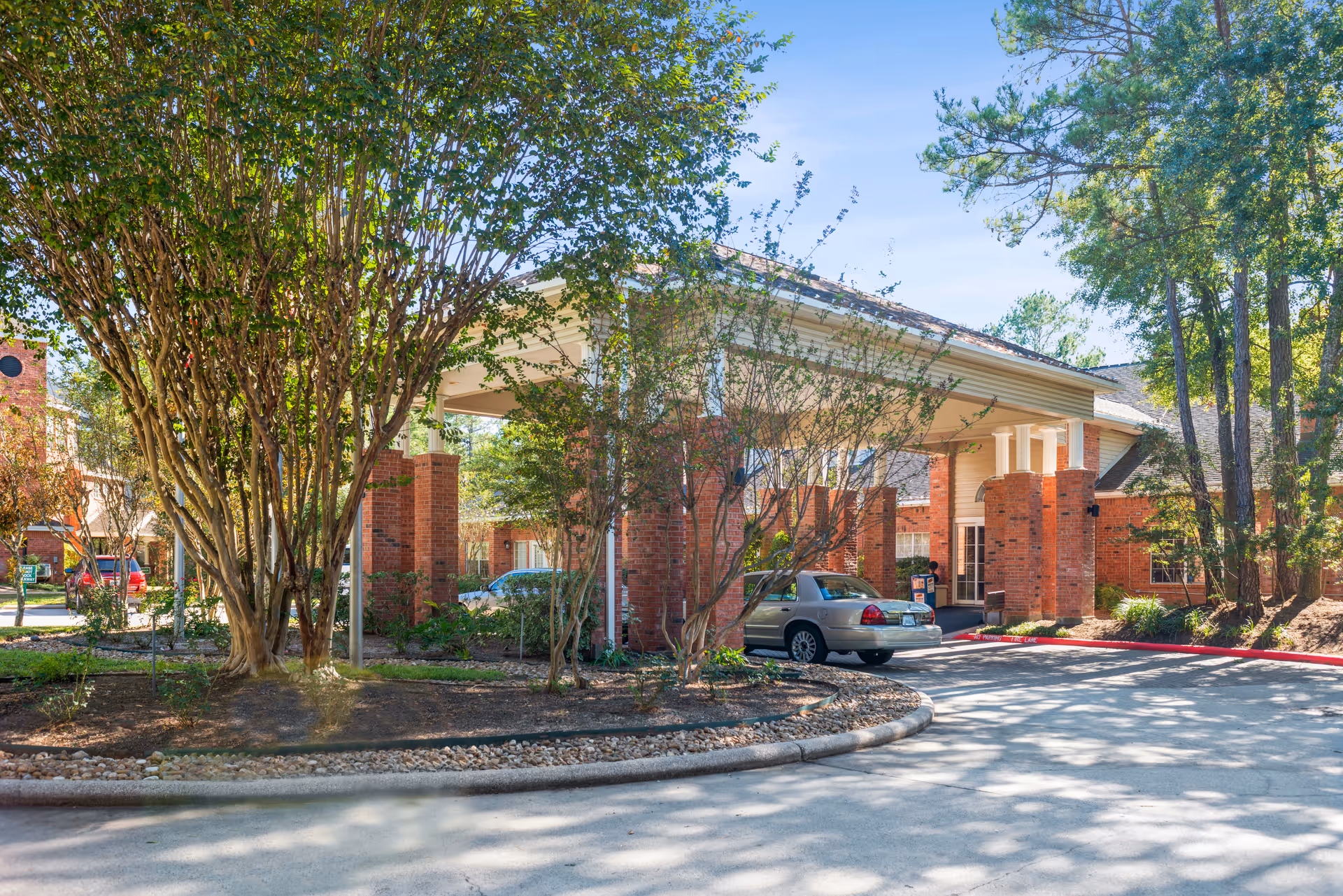 Exterior view of The Forum at The Woodlands senior living facility showing a covered entrance with brick pillars, surrounded by trees and landscaping. A silver car is parked near the entrance under a clear blue sky.