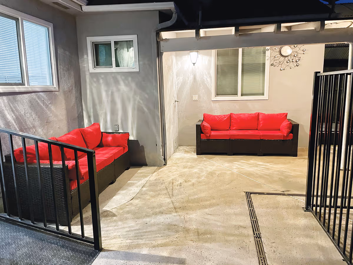 Covered outdoor patio with two red-cushioned wicker sofas against a building wall.