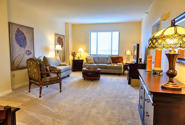 A well-lit living room with beige walls and carpet. The room features a large window with blinds, a beige sofa with cushions, a matching loveseat, and an upholstered armchair. There is a round ottoman in the center, two side tables with lamps, and a wooden dresser with a decorative lamp and mirror above it. Two framed floral artworks hang on the wall.