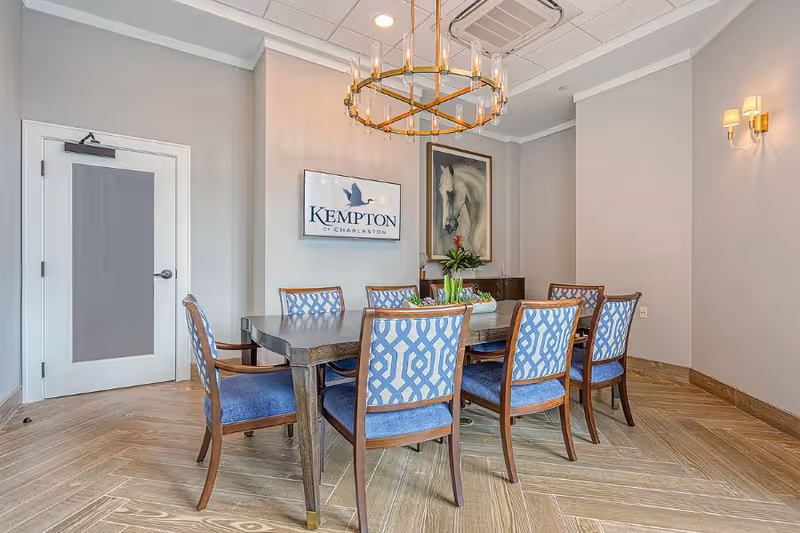 A dining room with a rectangular wooden table surrounded by eight blue cushioned chairs with patterned backs. On the wall, there is a framed sign that reads 'Kempton of Charleston' and a large painting of a white horse. The room has light-colored walls, a wooden floor with a herringbone pattern, a modern chandelier hanging above the table, and wall sconces providing additional lighting.