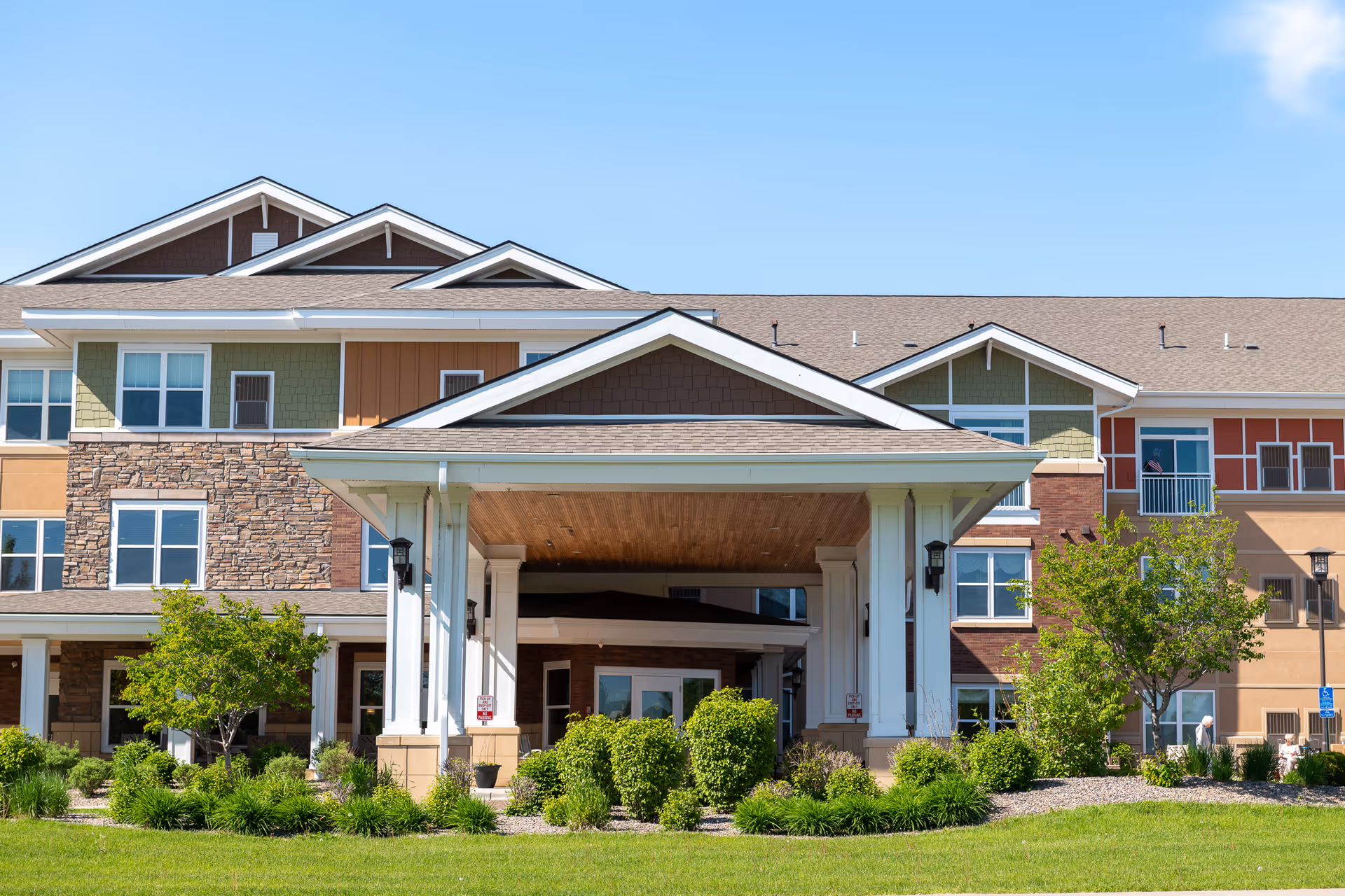 Front entrance of a multi-story senior living building with a covered porte-cochère, landscaping, and large windows.
