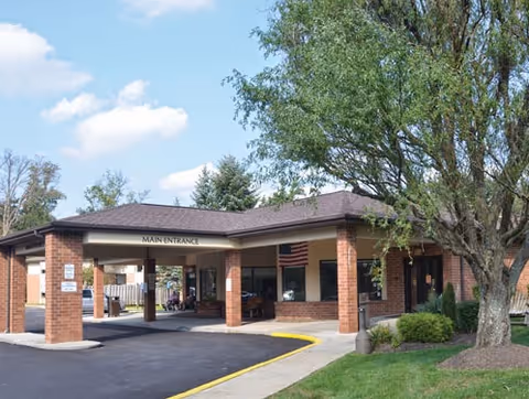 Exterior view of Amber Springs At Laurel Wood facility showing the main entrance with a covered drop-off area supported by brick columns. There is a paved driveway and sidewalk leading to the entrance, an American flag hanging near the entrance, and a large tree with green foliage on the right side. The sky is partly cloudy.