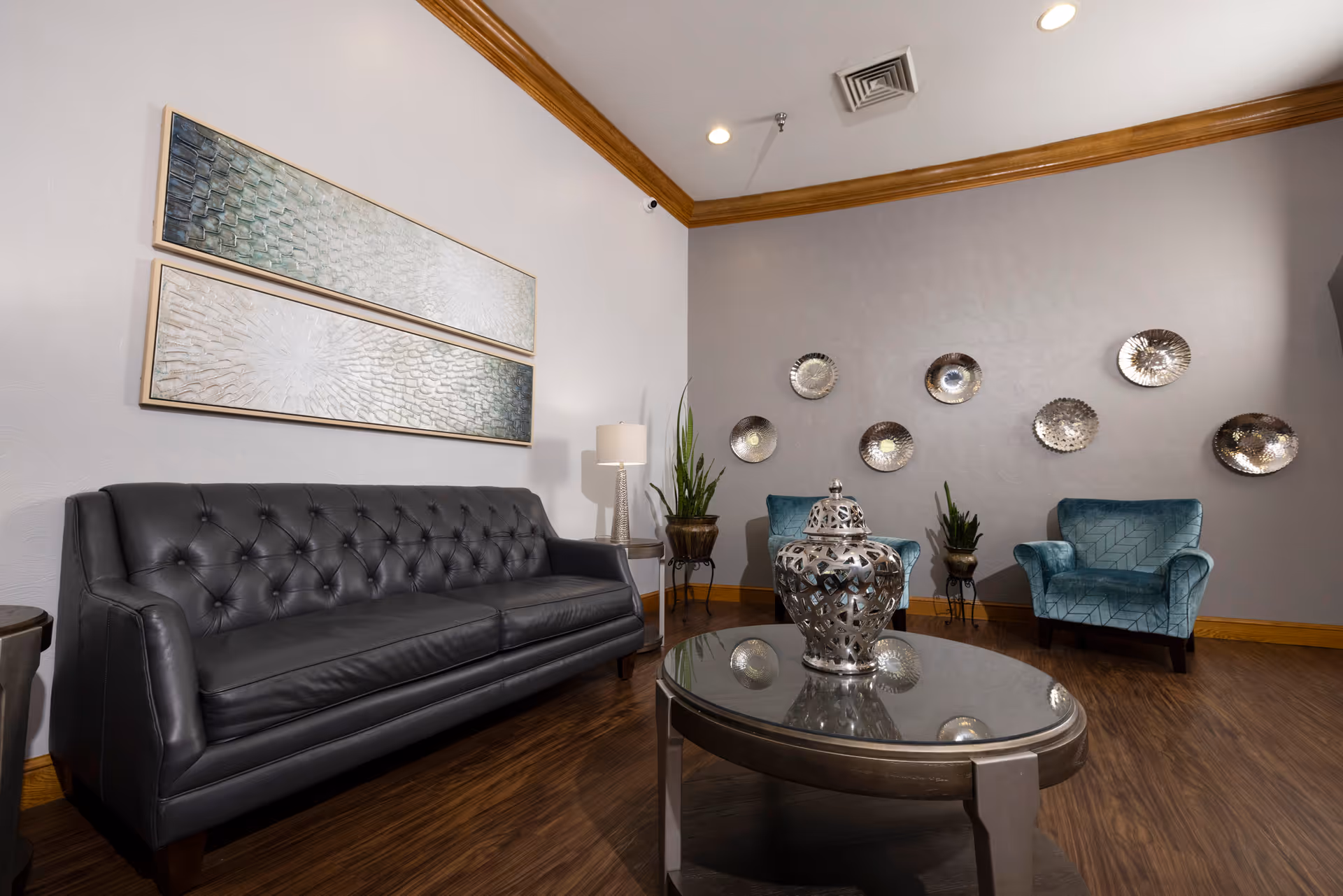 A cozy living room area with a black leather tufted sofa on the left, two teal armchairs on the right, and a round glass coffee table with a decorative silver vase in the center. The walls are decorated with two rectangular textured art pieces above the sofa and a collection of round metallic wall hangings on the adjacent wall. There are also two potted plants and a floor lamp with a white shade. The room has wooden flooring and crown molding along the ceiling.