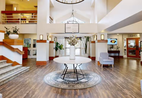 Bright, open lobby with a round table and floral arrangement on a rug, seating areas, staircase, and high ceilings.
