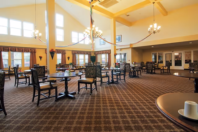 Spacious dining room in a senior living facility with multiple round tables and chairs arranged neatly on a patterned carpet. Large windows with curtains allow natural light to fill the room, and chandeliers hang from the high ceiling. The room has a warm and inviting atmosphere.