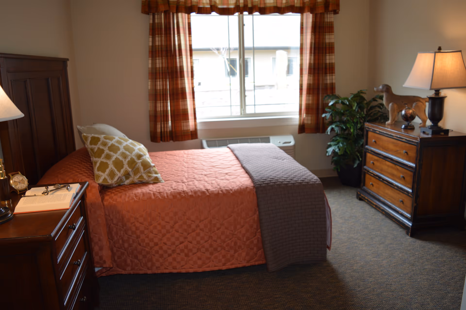 A cozy bedroom with a single bed covered in a rust-colored quilt and a gray throw blanket. There are two pillows on the bed, one with a yellow and white pattern. A wooden nightstand with a lamp, an alarm clock, and an open book is beside the bed. Across from the bed is a wooden dresser with a lamp, a decorative dog figurine, and a small globe. A window with plaid curtains lets in natural light, and a green potted plant is in the corner.