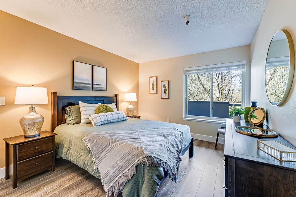 Sunlit furnished bedroom with a made bed flanked by nightstands and lamps, a dresser with round mirror, and a window overlooking trees.