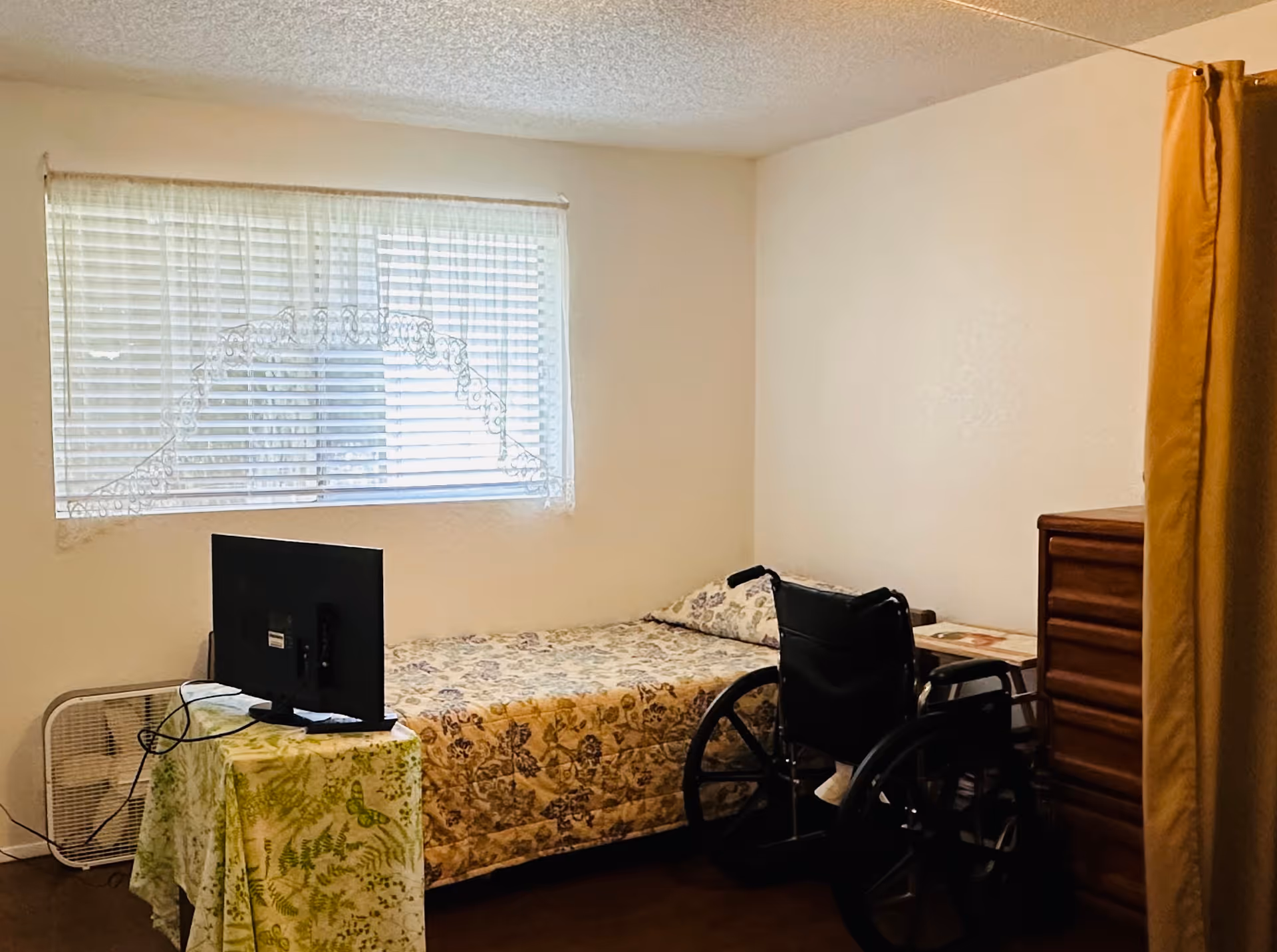 Small bedroom with a single bed, wheelchair, TV on a covered table, dresser, and a window with blinds.