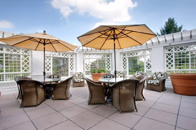 Outdoor patio area with two round glass tables, each surrounded by four wicker chairs with cushions. Two large beige umbrellas provide shade. There are additional cushioned wicker armchairs and large potted plants along the white lattice walls under a partly cloudy sky.