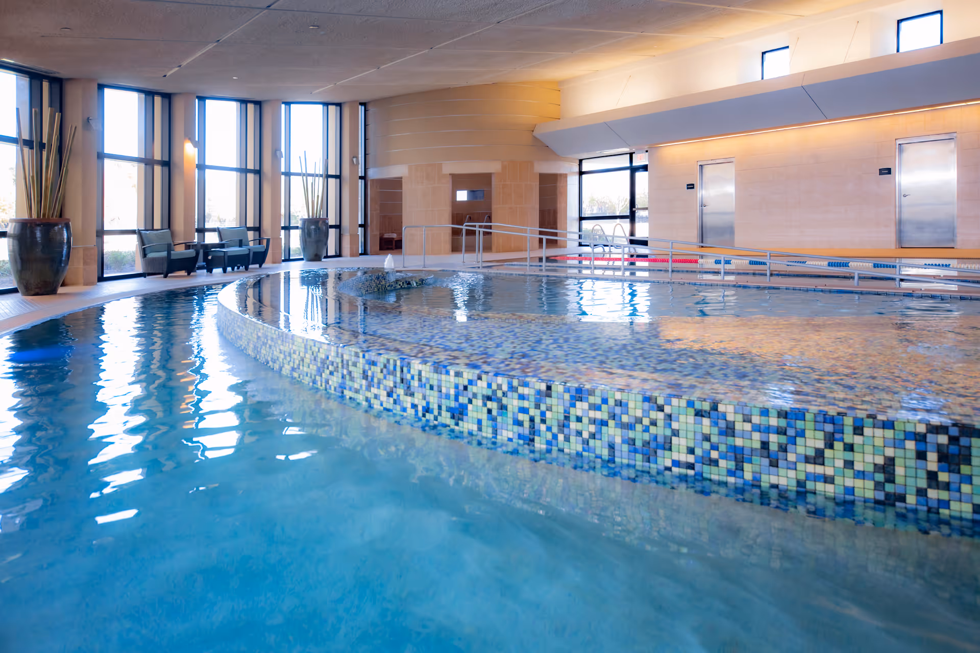 Indoor swimming pool with mosaic tile edge, surrounded by large windows letting in natural light. There are chairs and large potted plants along the windows, and a ramp leading into the pool area.