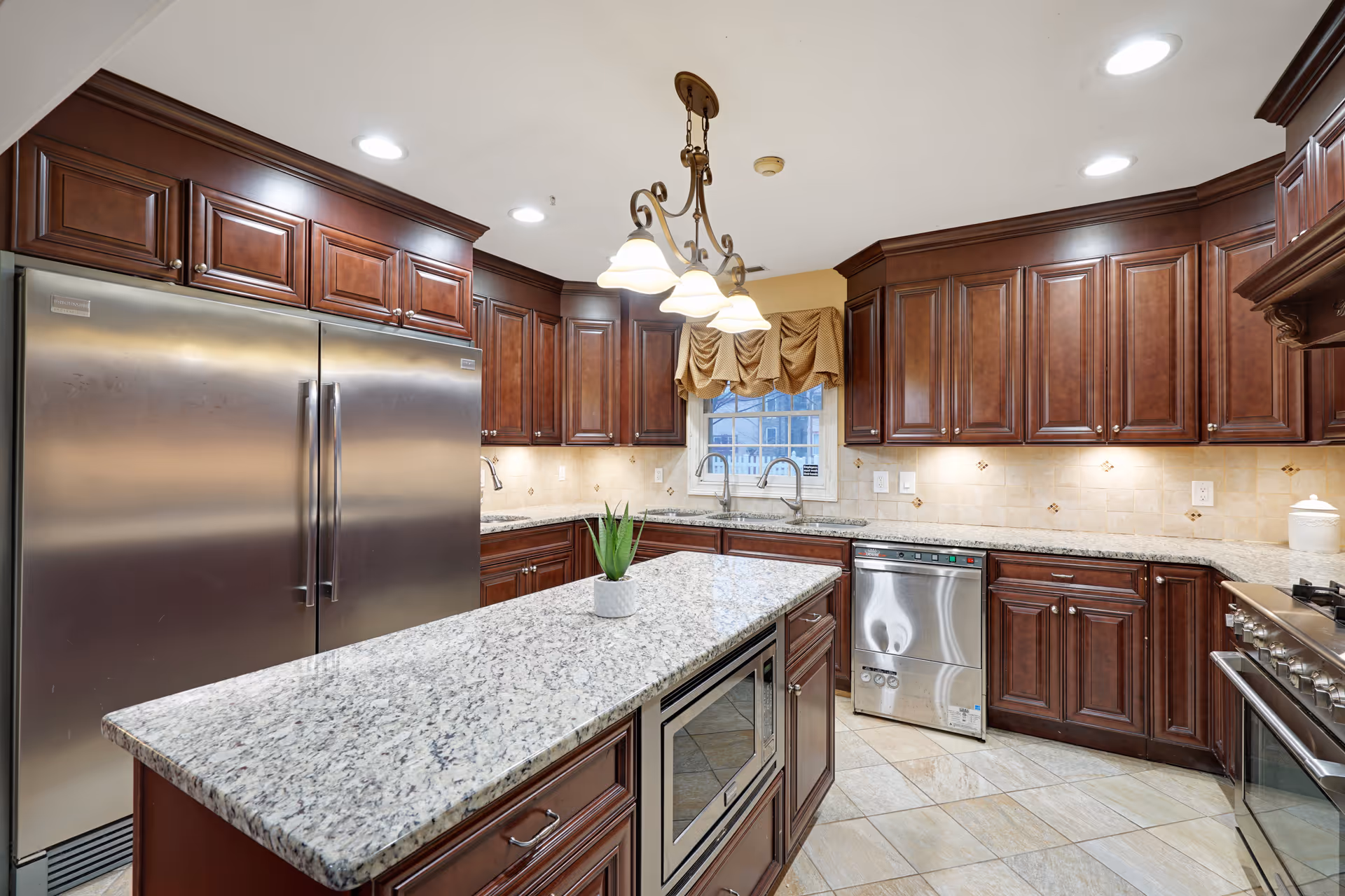 A spacious kitchen with dark wooden cabinets, granite countertops, and stainless steel appliances including a double-door refrigerator, dishwasher, and oven. There is a kitchen island with a built-in microwave and a small potted plant on top. A window with a beige valance is above the sink, and a decorative light fixture hangs from the ceiling.